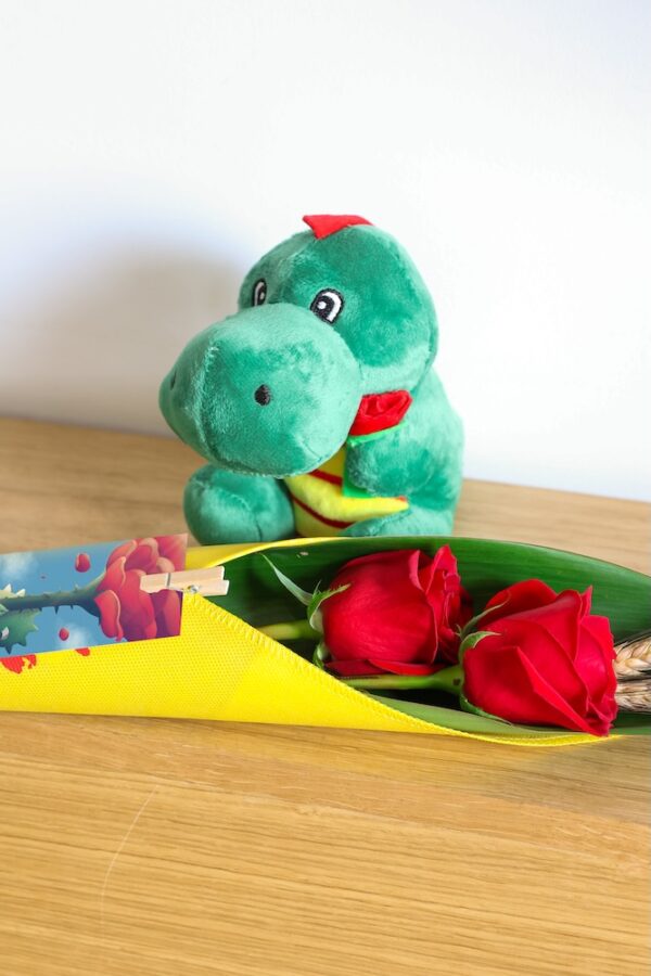 Cono de tela con 2 rosas rojas y espigas junto a peluche de dragón y punto de libro.