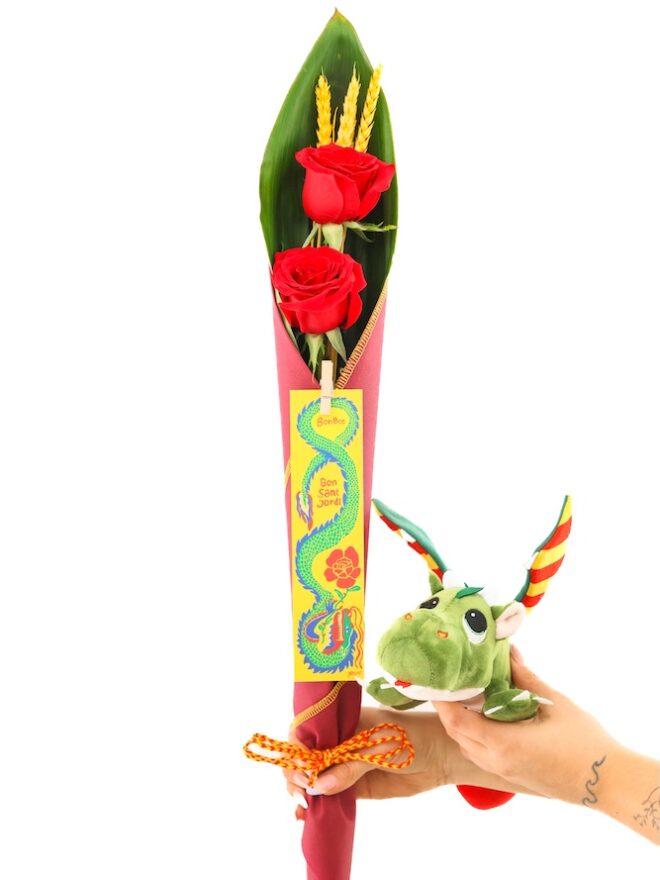 Cono de tela con 2 rosas rojas y espigas junto a peluche de dragón y punto de libro.